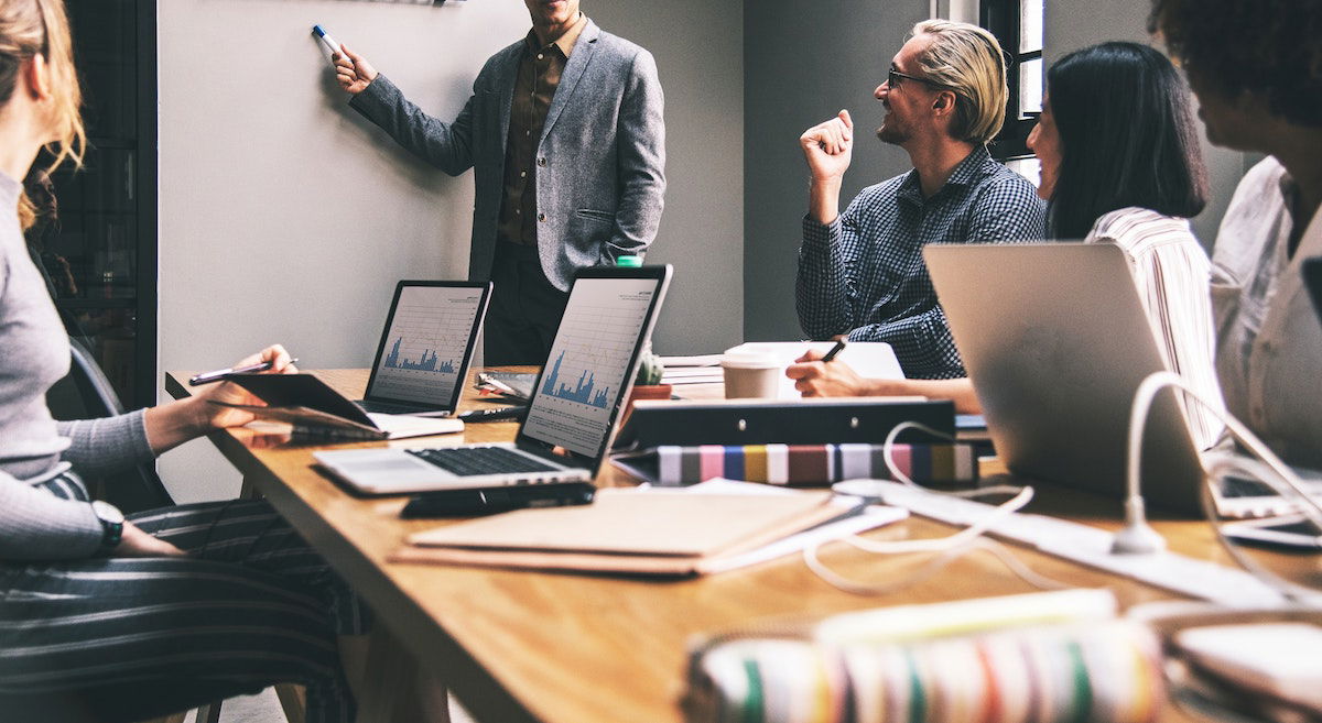 A group of people in an office setting sit around a table with laptops, notebooks, and documents. One person stands at a whiteboard, presenting. Others listen attentively. The atmosphere suggests a professional meeting or discussion.