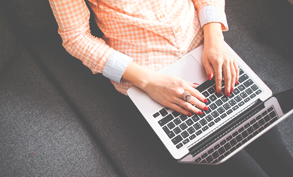 Person typing on a laptop while sitting on a gray couch. They are wearing a light orange checkered shirt and have red nail polish.