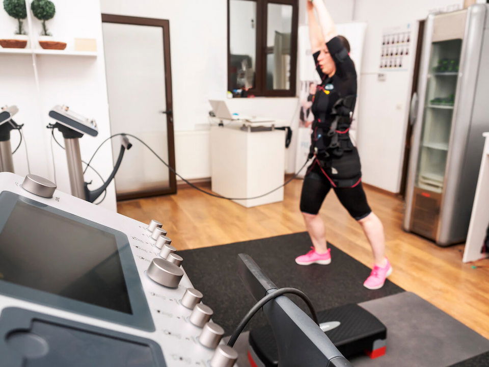 A person wearing exercise gear performs a workout in a fitness studio equipped with various electronic devices and machines. The room has wooden flooring and a mix of modern equipment, with a white wall and closed door in the background.