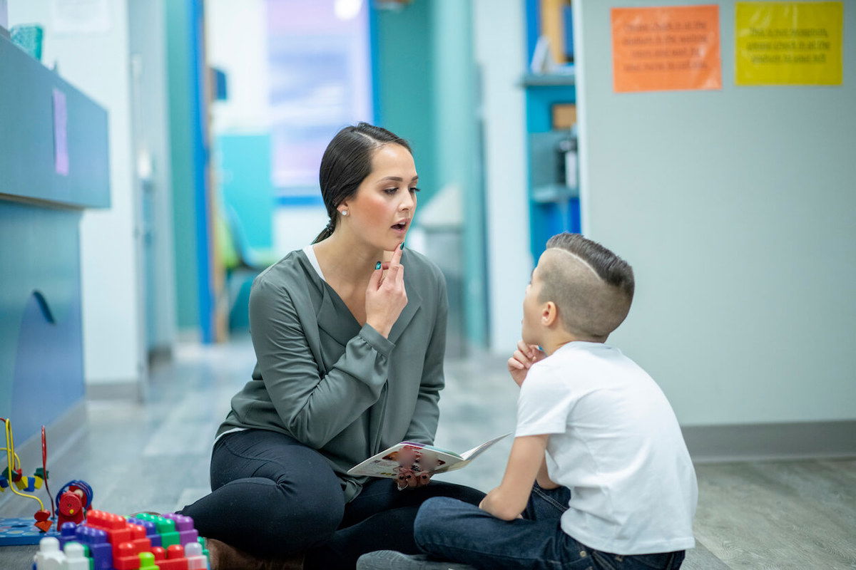 A woman sits on the floor with a young boy in a therapy setting. She holds a mirror and points to her mouth, likely demonstrating speech sounds. The boy watches attentively. Colorful toys are visible on the floor nearby.