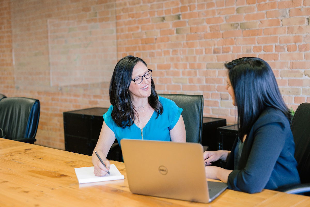 Two women are sitting at a wooden conference table with a laptop open. They are engaged in conversation, with one taking notes. Both are smiling, and the room has exposed brick walls and black chairs.