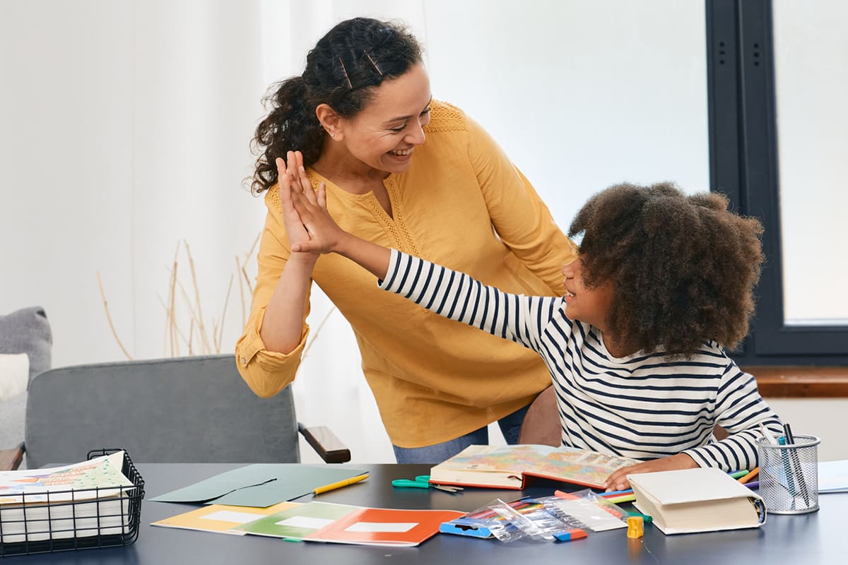 A woman wearing a yellow shirt and a child in a striped shirt share a high five at a table with colorful papers and notebooks. They appear happy, engaged in an activity together in a bright room.