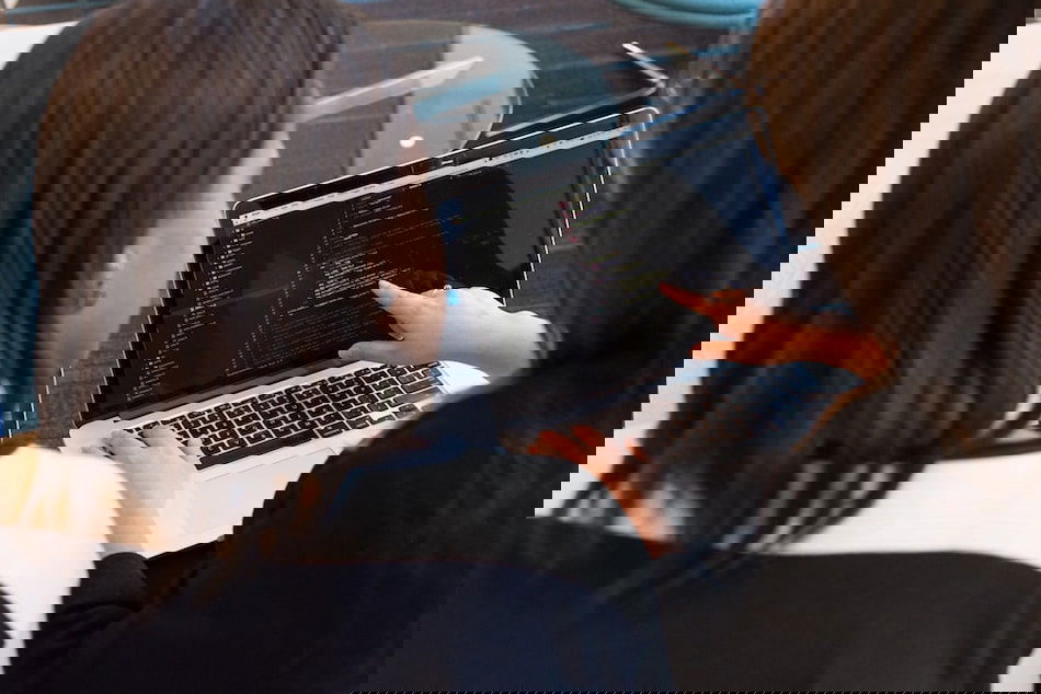 Two people sitting at a table look at a laptop screen displaying code. One person points at the screen while the other observes. They are discussing the content on the laptop. The background is an office setting with glass and modern furniture.