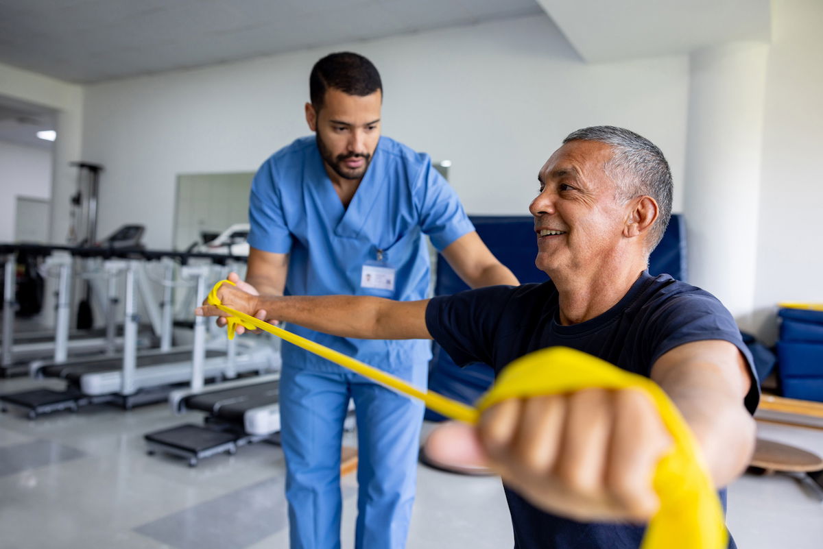 A healthcare professional assists an older man with resistance band exercises in a physical therapy setting. The man smiles as he stretches the yellow band, while the therapist, in blue scrubs, supervises. Exercise equipment is visible in the background.