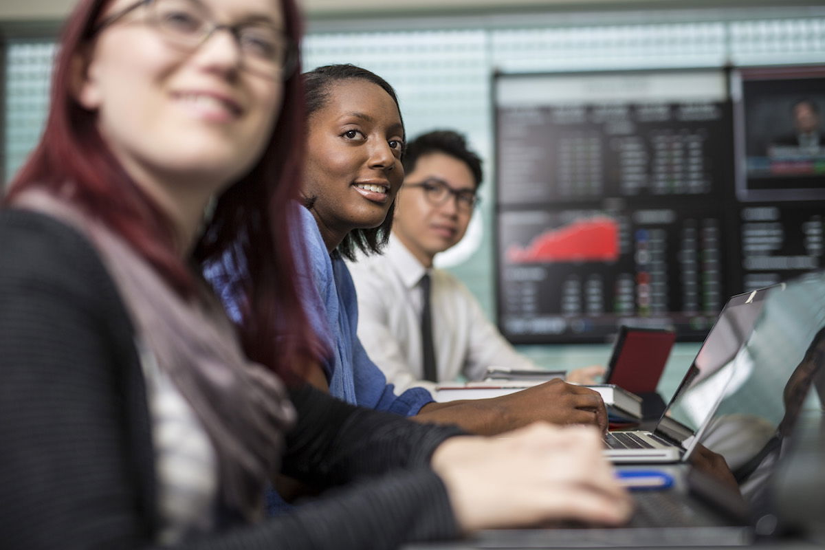 A group of three people sitting in a conference room, using laptops. In the background, multiple monitors display financial data and graphs. They appear focused and engaged in their work.
