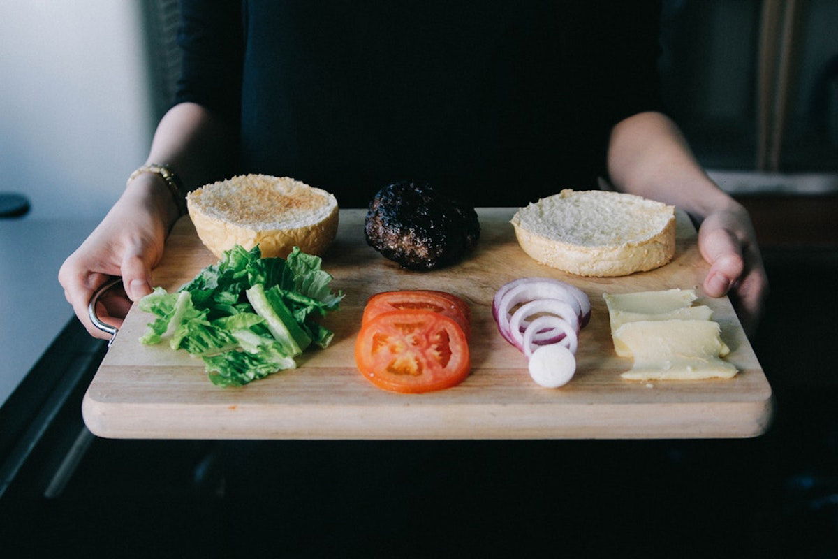 A person holds a wooden board with ingredients for a burger: a bun, a cooked patty, lettuce, tomato slices, onion rings, and cheese slices, all arranged in a row. The person wears a black top, with only their arms visible.