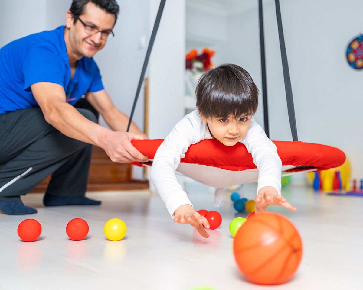 A young child in a white shirt is lying on a red swing, reaching for colorful balls on the floor. An adult in a blue shirt is assisting and watching them in a playful indoor setting.