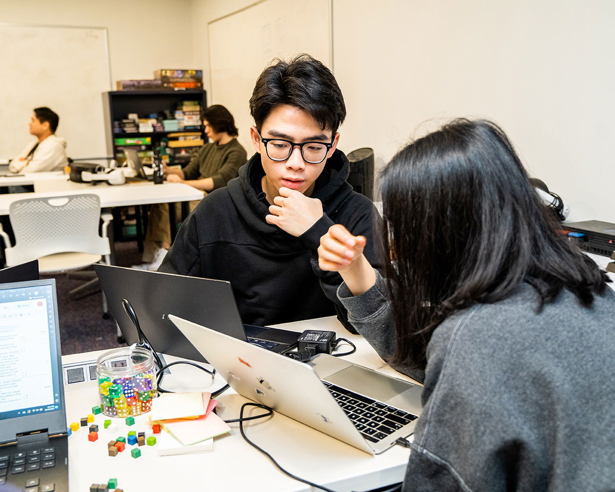 Two people sit at a table, each with a laptop. They are engaged in conversation, with one person gesturing. The table is covered with laptops, a notebook, and colorful dice. Other people work in the background.