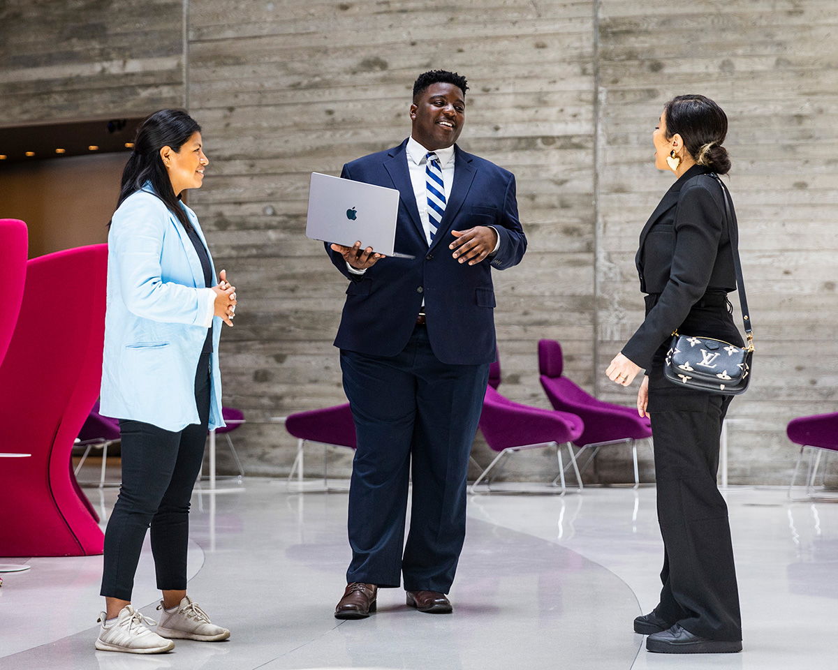 Three people in business attire stand in conversation in a modern office space with pink chairs and a concrete wall. One person holds a laptop. They appear engaged and friendly.