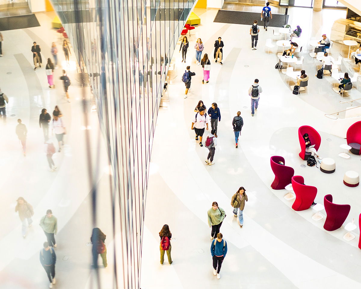 A large, bright atrium with people walking and talking. On the right, there's seating with red chairs and white tables. The left side features a reflective glass wall, creating a mirrored effect of the space and people.