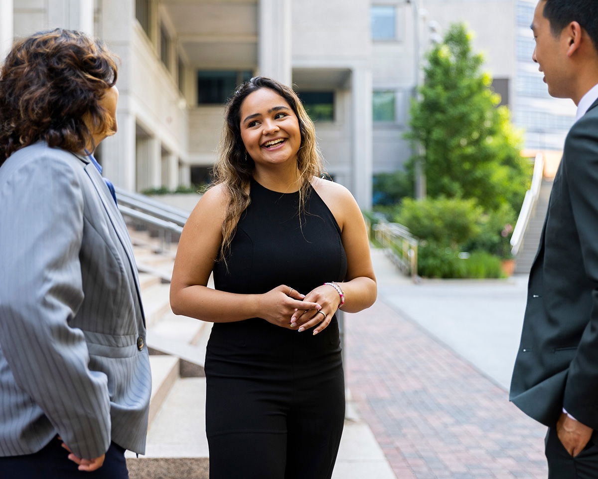 Three people are standing on a city street, engaged in conversation. The woman in the center is smiling, wearing a black sleeveless top. They are near a building with trees and steps in the background.