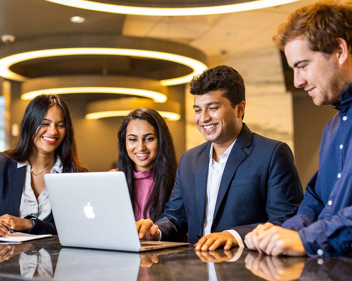 A group of four people gathered around a laptop, smiling and engaged. They are seated at a table with circular ceiling lights in the background, suggesting a modern office or collaborative workspace.