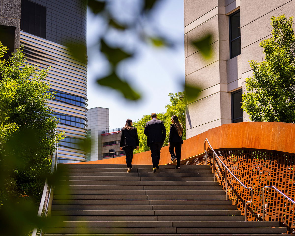 Three people in suits walking up outdoor stairs surrounded by greenery and modern buildings. The scene suggests an urban setting with a mix of nature and architecture.