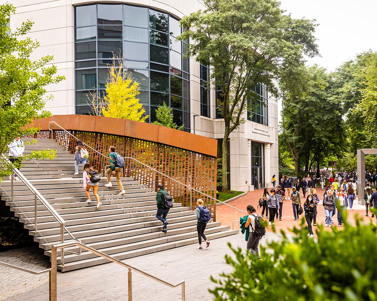 A group of students walks up and down a wide staircase outside a modern university building with large windows. The scene is bustling with activity, surrounded by lush green trees and pathways.