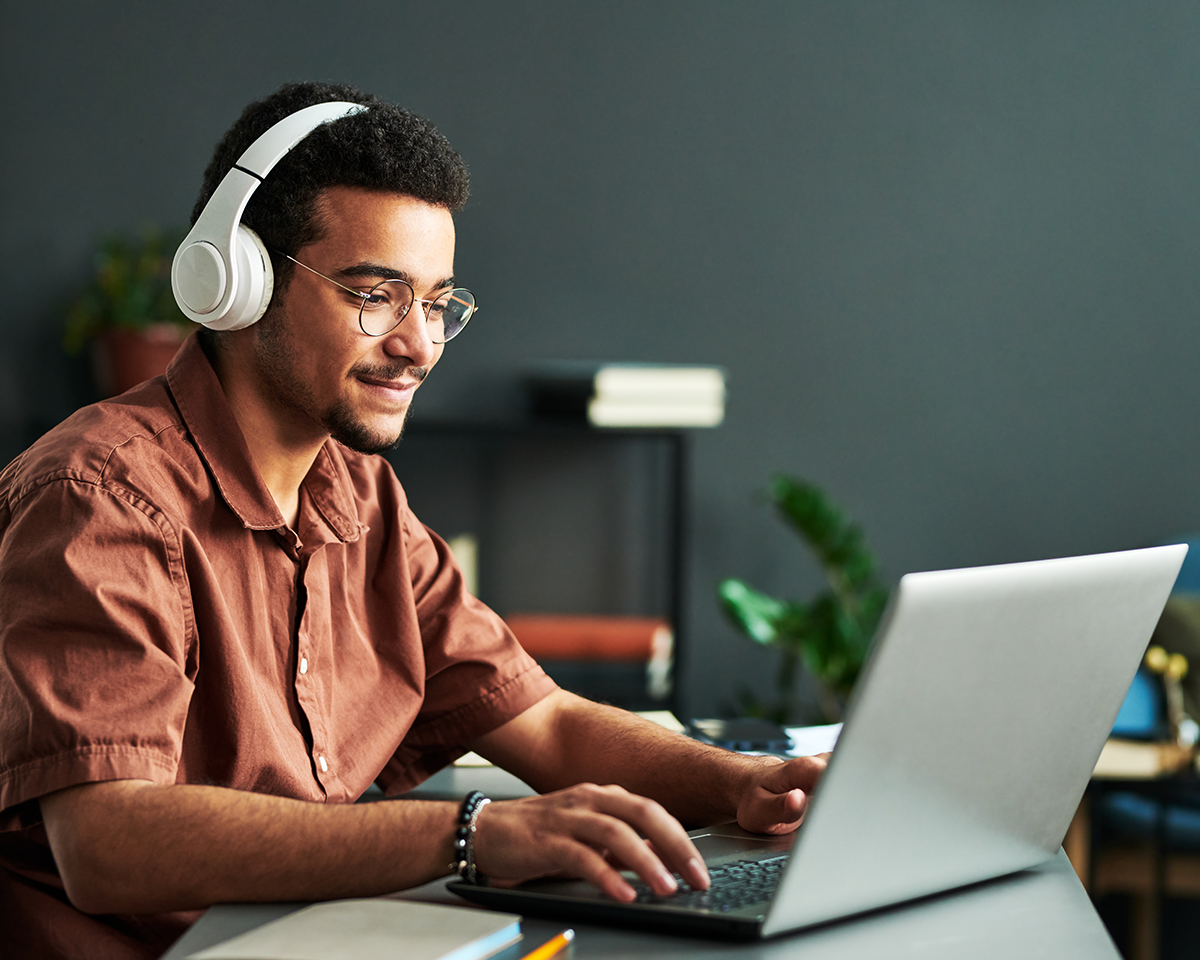 A person wearing headphones and glasses is sitting at a desk, using a laptop. They are smiling and appear focused. The background includes a dark wall and some blurred books and plants.