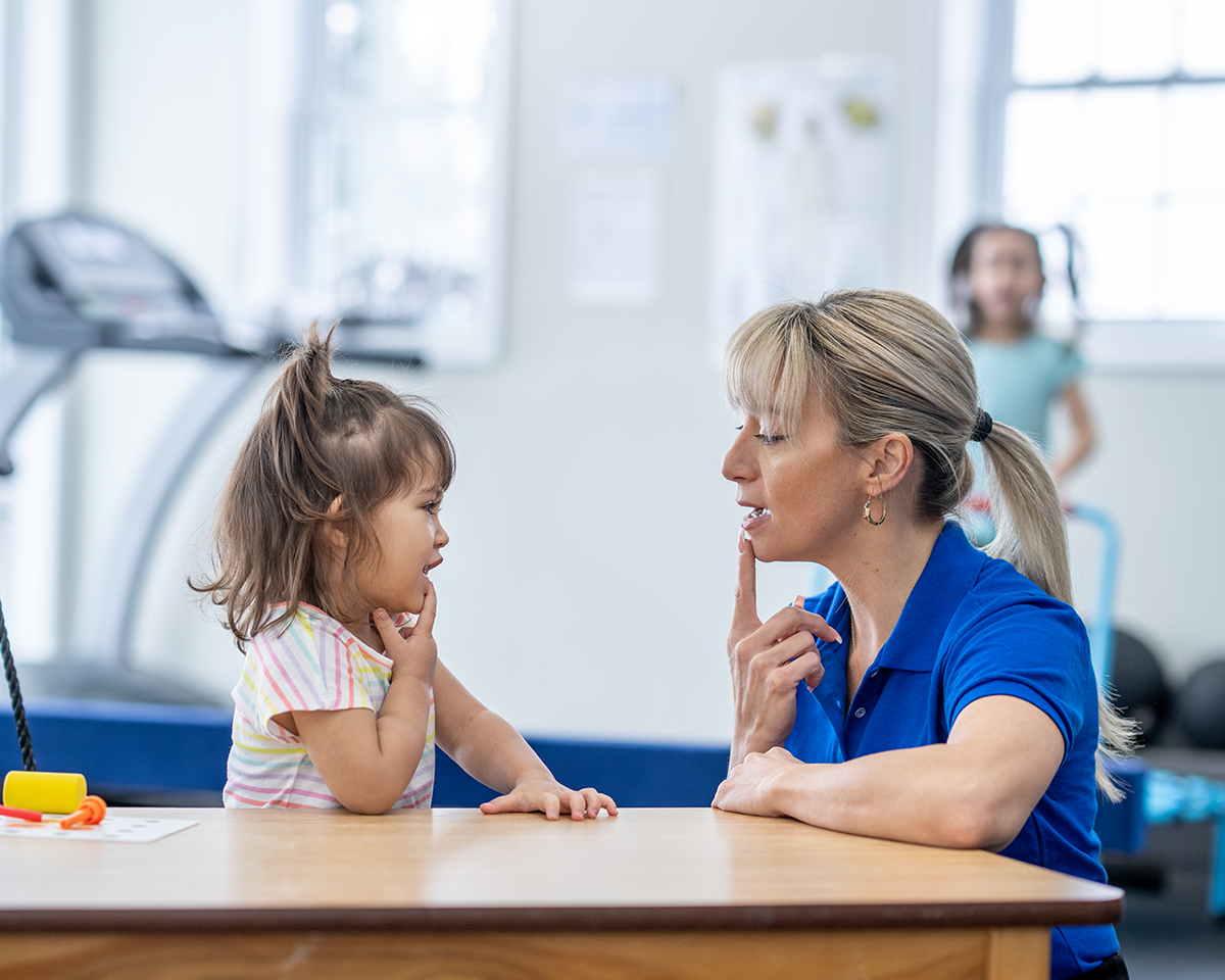 A young girl and an adult woman sitting at a table, both touching their chins with their fingers, seemingly engaged in a learning or speech therapy activity. The background shows a treadmill and gym equipment.