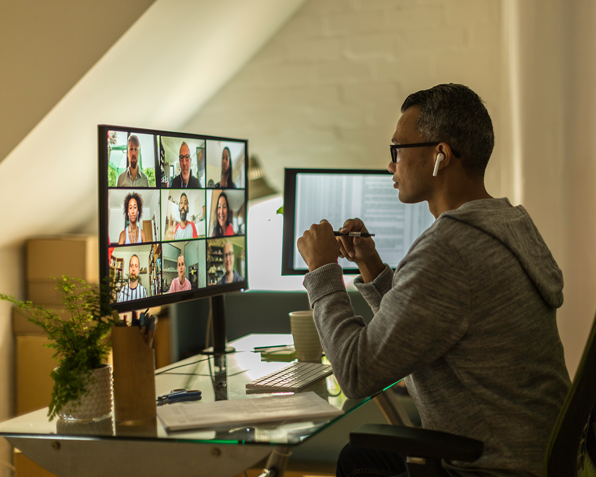 A person in a gray hoodie sits at a desk, using a computer to participate in a video conference with multiple people. The desk has two monitors, a potted plant, a notebook, and a coffee cup. The scene is set in an attic office space.