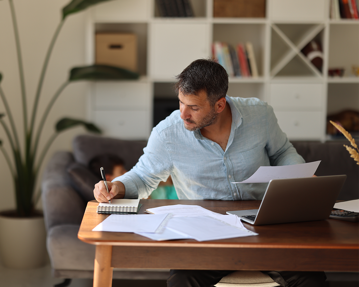 A man in a light blue shirt sits at a wooden table, working on papers and using a laptop. A notebook and sheets of paper are spread out before him. The background features a couch and a bookshelf with decorative items.