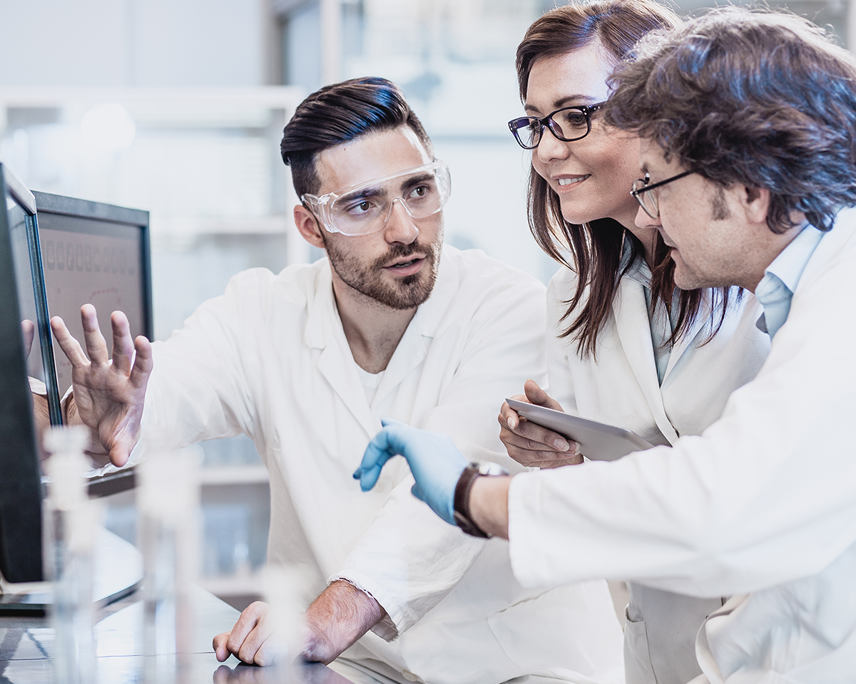 Three scientists in lab coats and safety glasses discuss research findings on a computer screen. One is gesturing towards the screen, while another holds a tablet. Lab equipment is visible on the table.