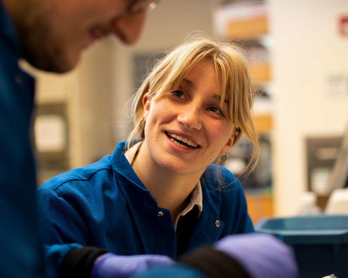 A woman in a blue lab coat smiles and talks to a man in the foreground. She has blonde hair tied back, and both are engaged in a laboratory. The scene is well-lit, and there are lab equipment and shelves in the background.