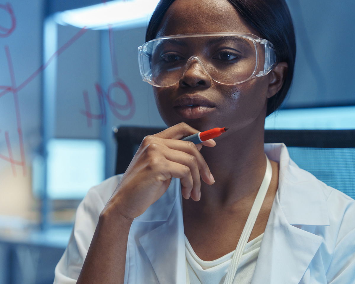 A person wearing safety goggles and a lab coat holds a marker while looking thoughtfully at chemical formulas drawn on a glass board in a laboratory setting.