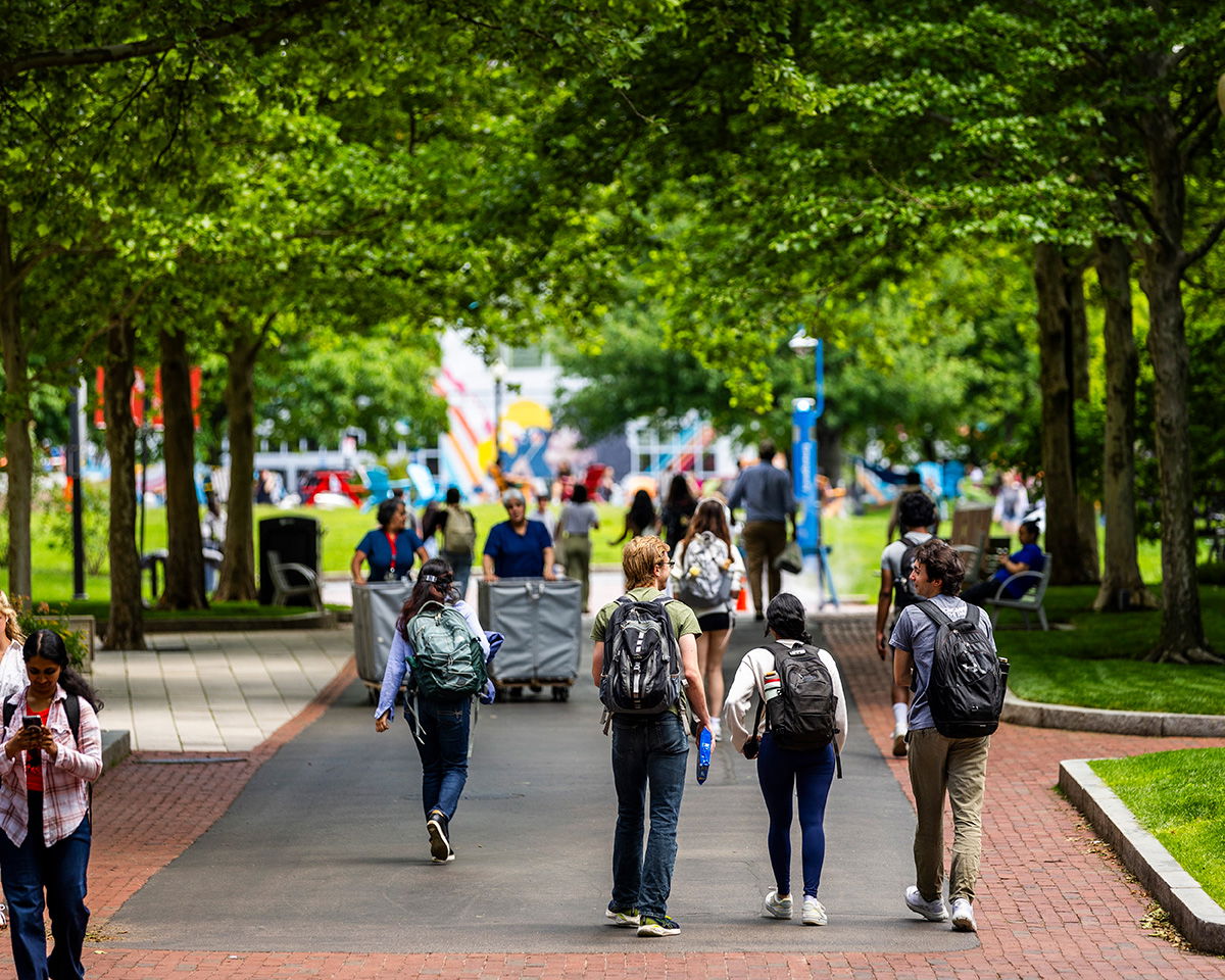 A group of students with backpacks walk along a tree-lined path on a campus, surrounded by green grass and shaded by large trees. Other people are seen in the background near benches and colorful flags.