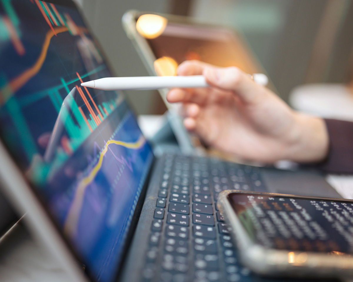 A hand holding a stylus is pointing at a financial graph on a tablet screen. The tablet is connected to a keyboard, and a smartphone with text is visible in the foreground. The graph shows fluctuating data in red and green lines.