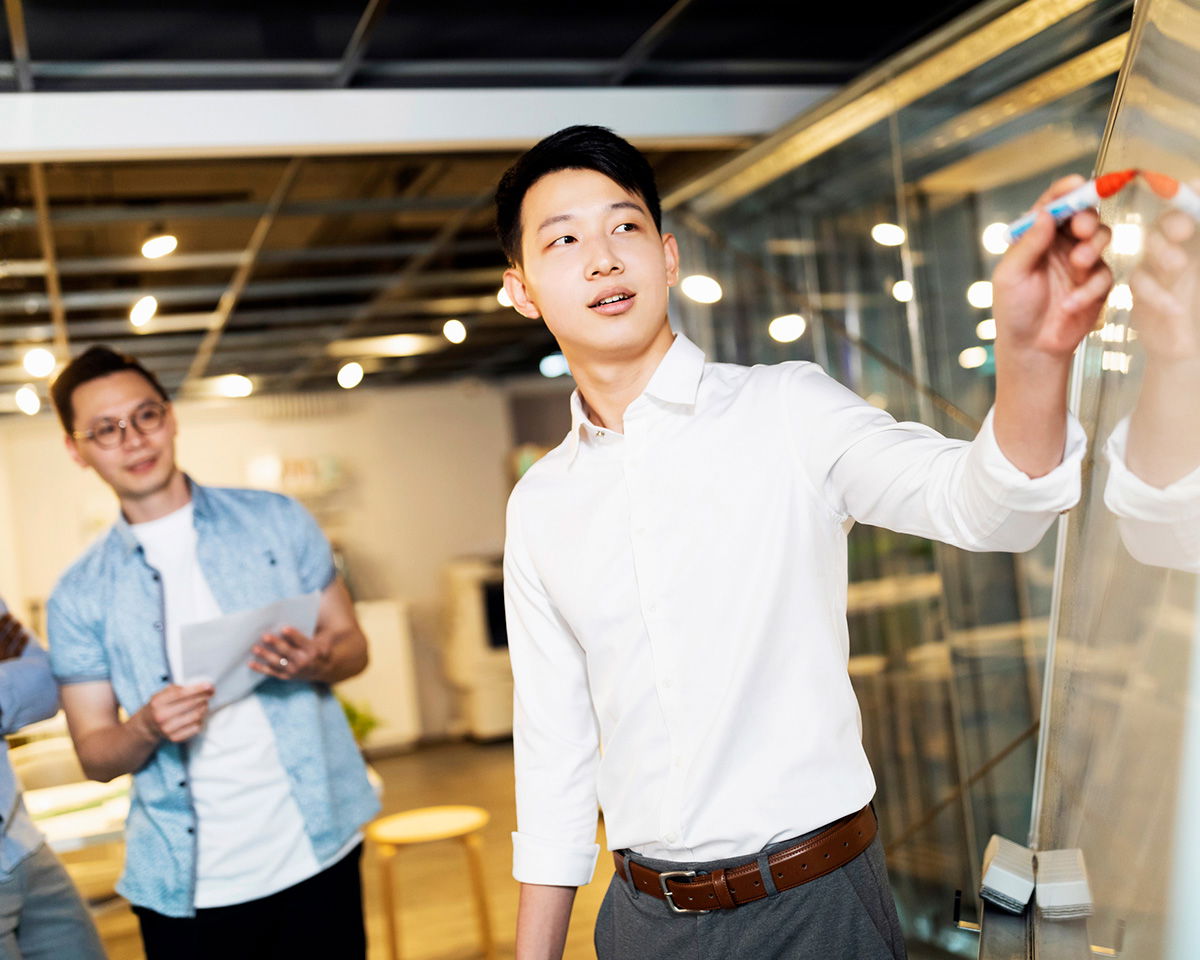 A man in a white shirt writes on a glass board with a marker while a group of people, including a man in a blue patterned shirt holding papers, looks on. The setting appears to be a modern office space with bright lighting and open design.