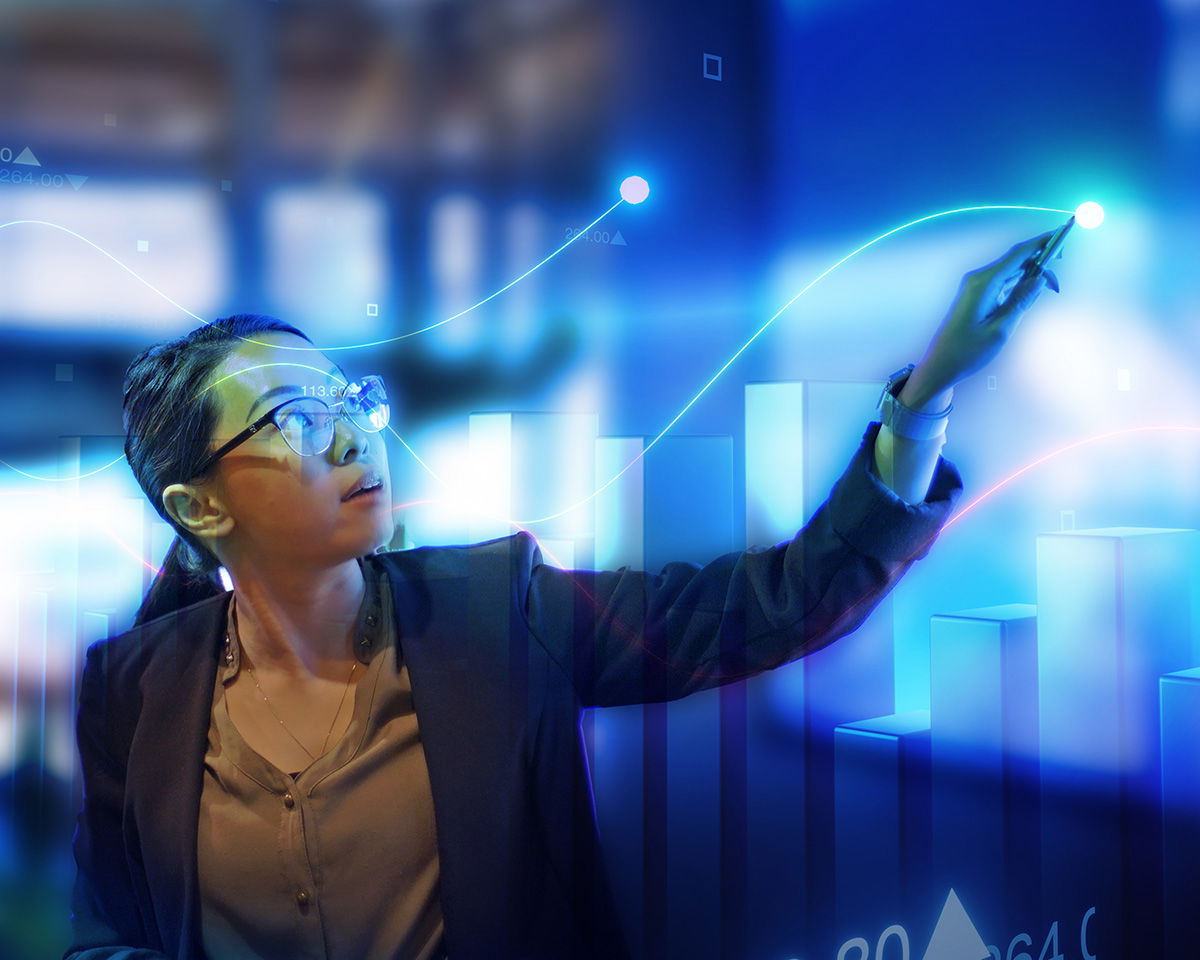 A person with glasses interacts with a large digital screen displaying graphs and bar charts. The environment is illuminated with blue tones, highlighting data points and analytics. The scene suggests a focus on technology and data analysis.