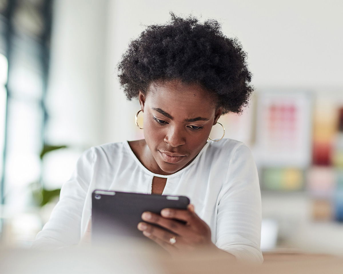 A person with curly hair, wearing a white blouse and hoop earrings, is intently looking at a tablet. The background is softly blurred, suggesting an indoor setting with natural light.