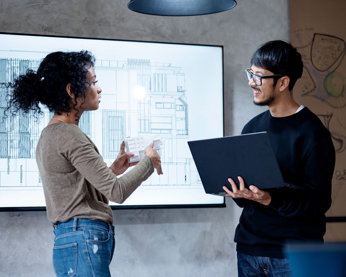 Two people are in an office setting discussing a blueprint displayed on a screen. One person holds a tablet and gestures, while the other holds a laptop and listens attentively. The background includes a modern design.