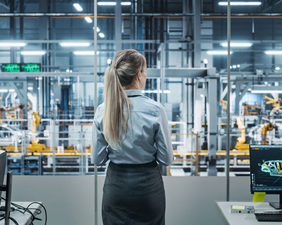 A woman with long hair, wearing a professional outfit, stands in a room overlooking an advanced factory floor. Robotics and machinery are visible through the large glass window. A computer screen displays technical information nearby.