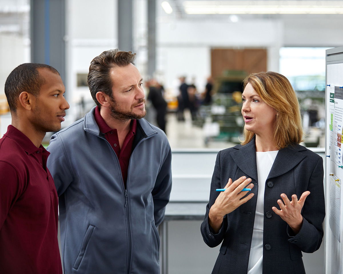 Three people are engaged in discussion in a modern workspace. One woman gestures while speaking to two men. The room is bright and open, with office equipment visible in the background.