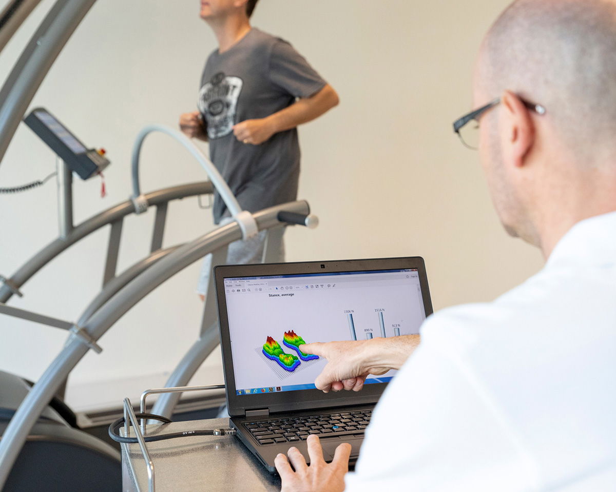 A man in a t-shirt runs on a treadmill while another man sits at a desk nearby, looking at a laptop. The laptop screen displays a colorful 3D graph and data charts. Exercise monitoring equipment is visible around the treadmill.