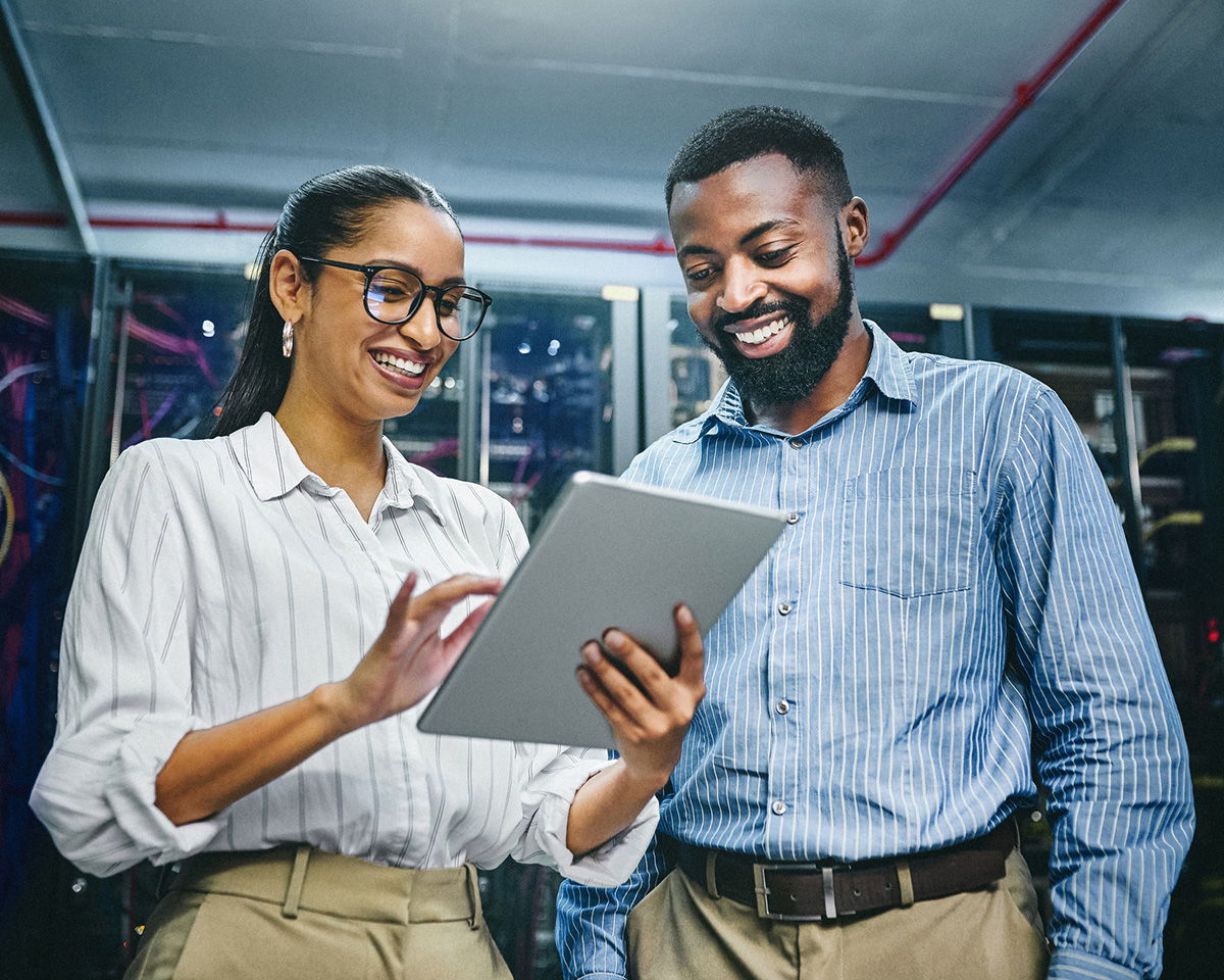 Two people stand in a server room, both smiling. The person on the left, wearing glasses and a striped shirt, holds a tablet. The person on the right, with a beard and denim shirt, looks at the screen. Server racks are visible in the background.