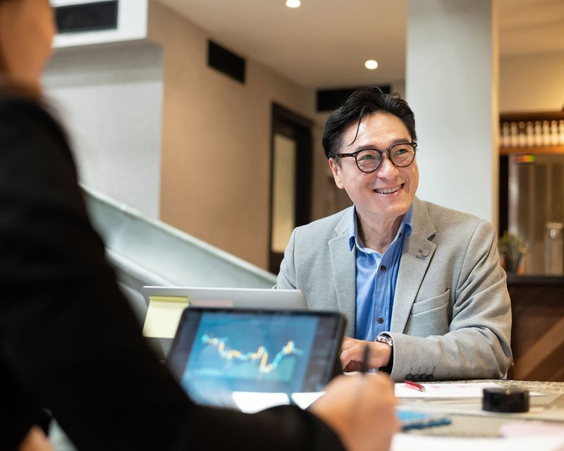 A man in a grey blazer and blue shirt sits at a table, smiling and engaged in conversation. A tablet on the table displays a graph. Another person is partially visible in the foreground. The setting appears to be a modern office or conference room.