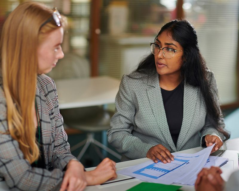 Two women in a meeting sit at a table with documents. One woman with long dark hair and glasses is speaking, while the other, with long blonde hair, listens attentively. They are in an office setting.