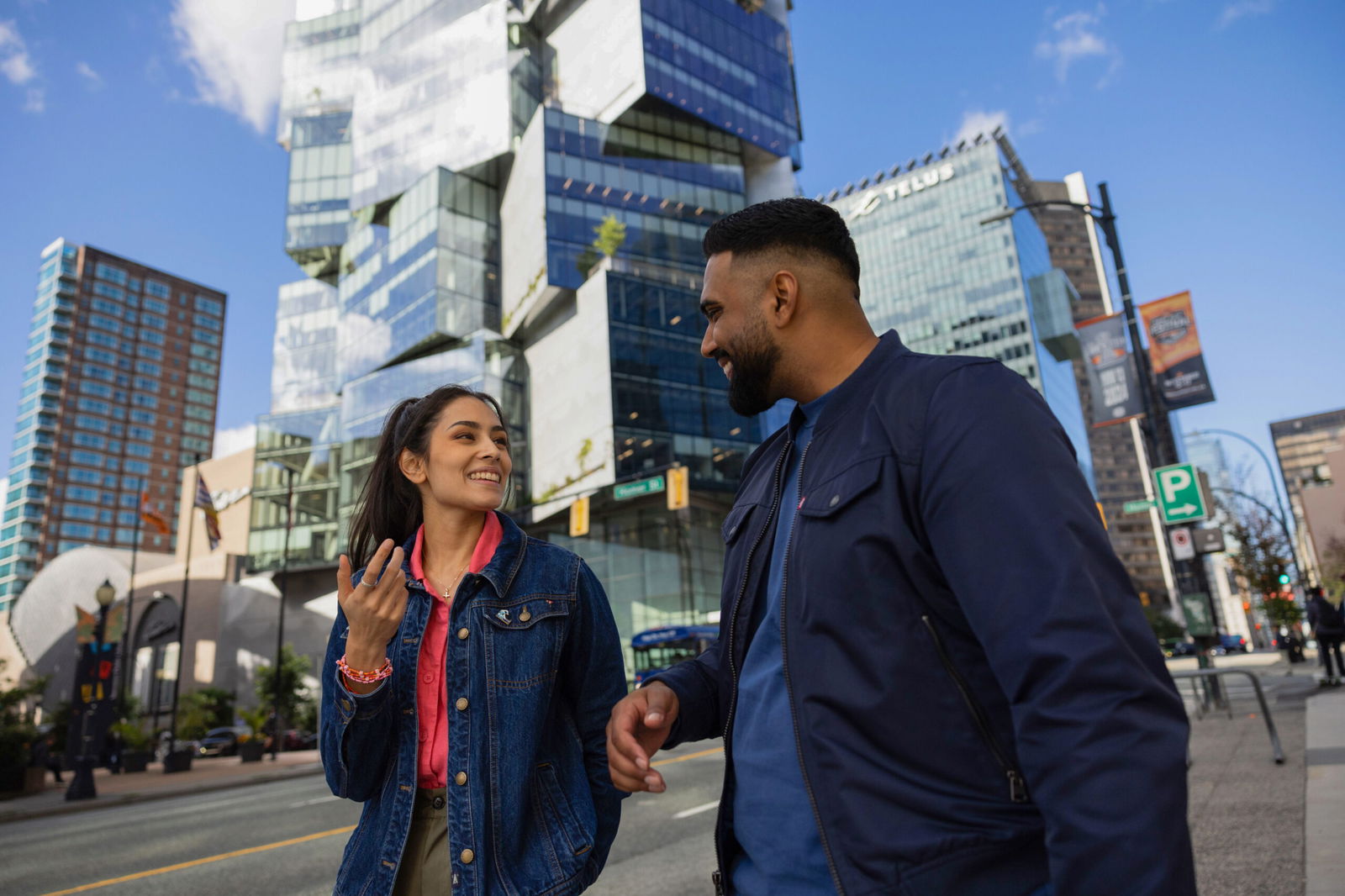 A man and a woman are walking and smiling on a city street with modern buildings in the background. The sky is clear and sunny, and they are dressed in casual jackets.