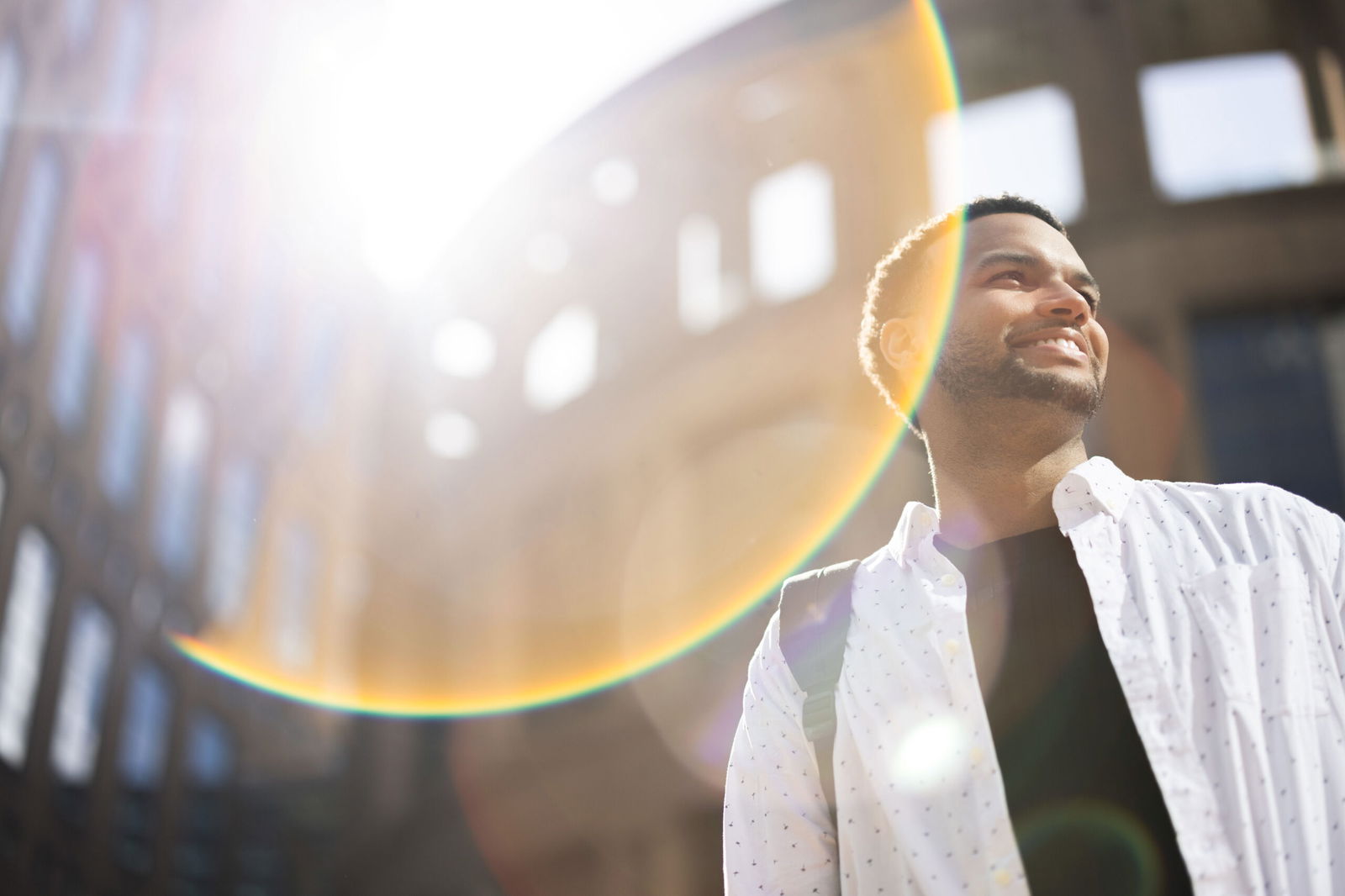 A man in a white shirt smiles while standing outdoors with a sunlit urban building in the background. Sunlight creates a lens flare, adding colorful arcs across the image, emphasizing a bright and joyful atmosphere.