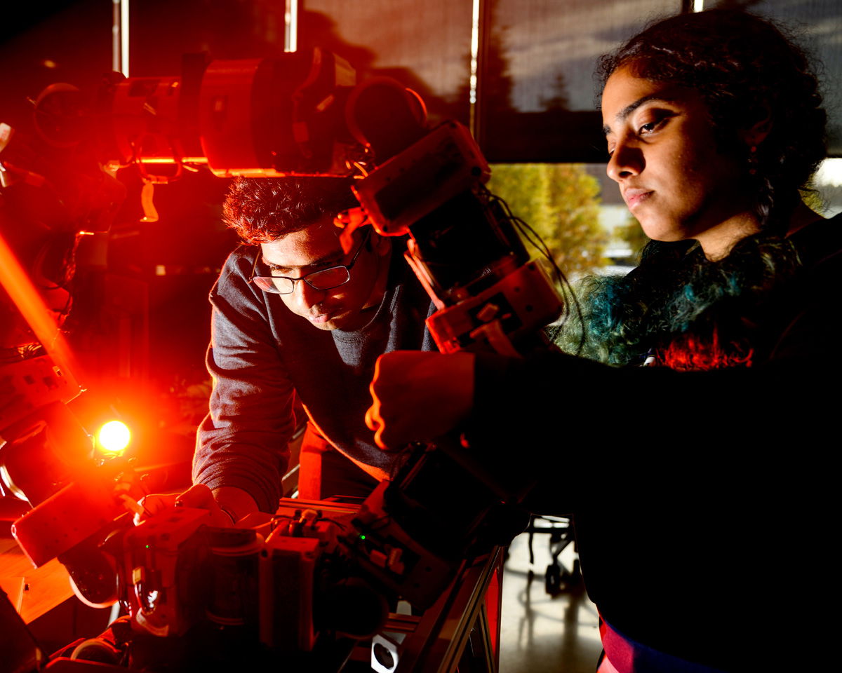 Two people work intently on a complex robotic arm in a dimly lit room. Red lighting accentuates the scene, highlighting the focus on the technology and the collaborative effort involved.