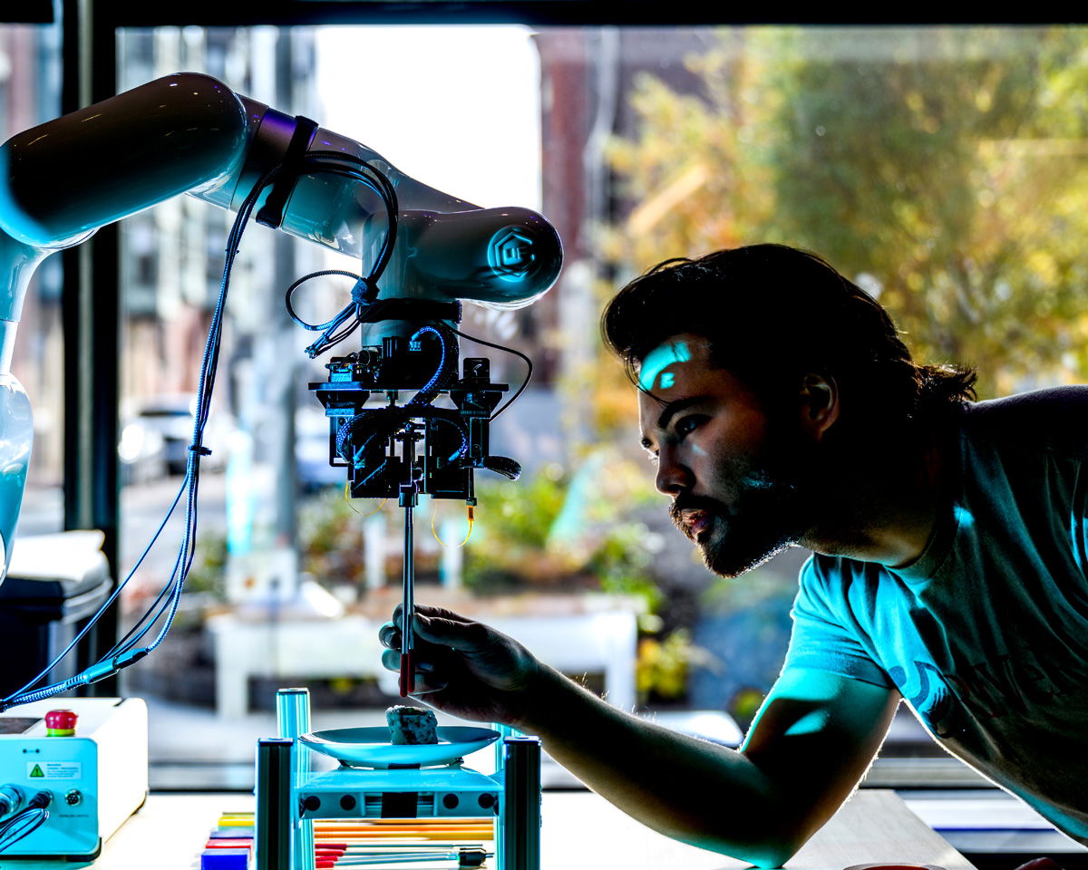 A person carefully adjusts a robotic arm in a bright, modern workspace. The setup includes a variety of colorful items on the table, with a window showcasing a blurred outdoor view in the background.